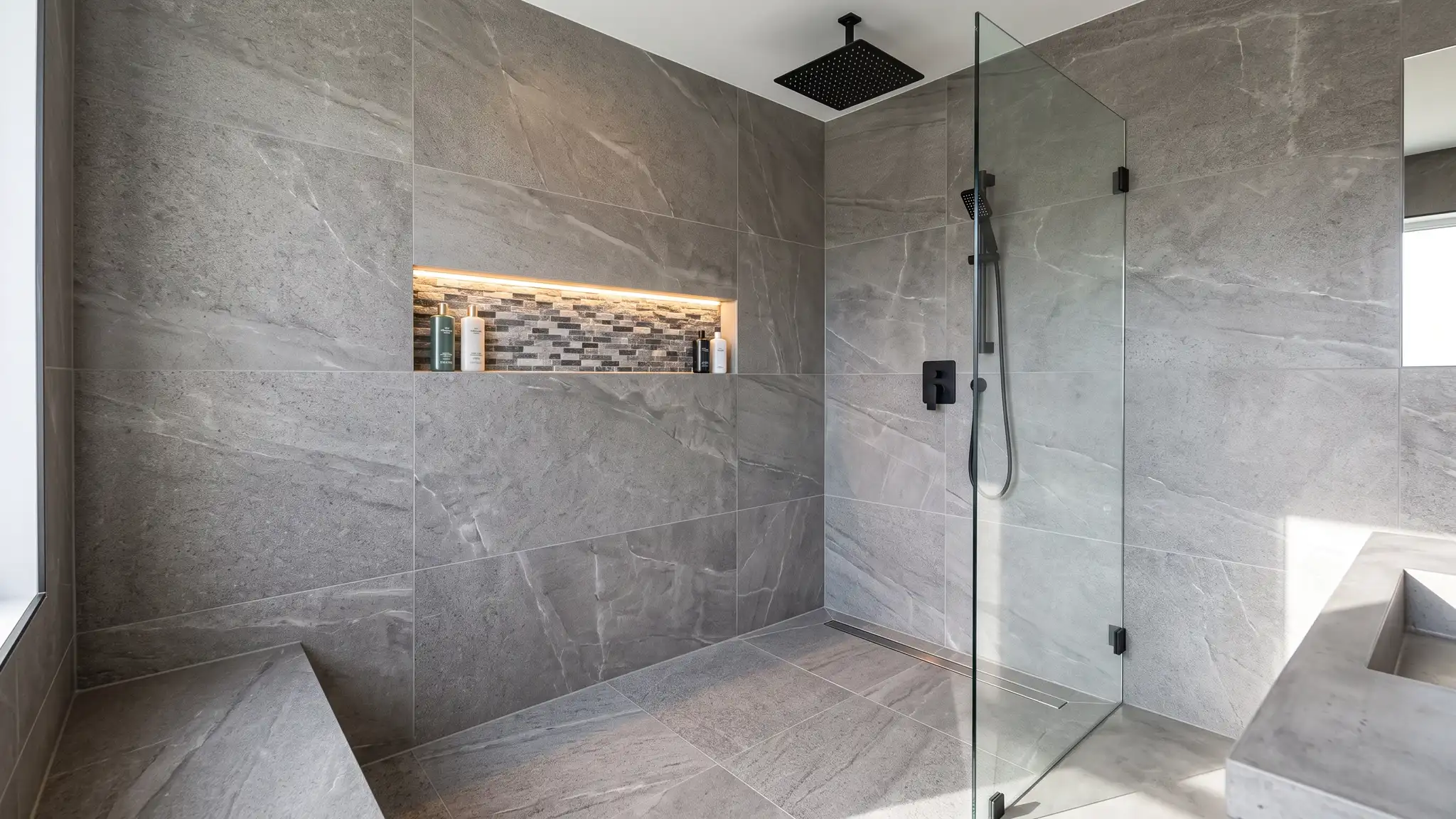 A beautiful floor-to-ceiling tiled walk-in shower with large format gray stone-look porcelain walls, a recessed niche with mosaic accent tile, a linear drain, a frameless glass panel and a rainfall shower head