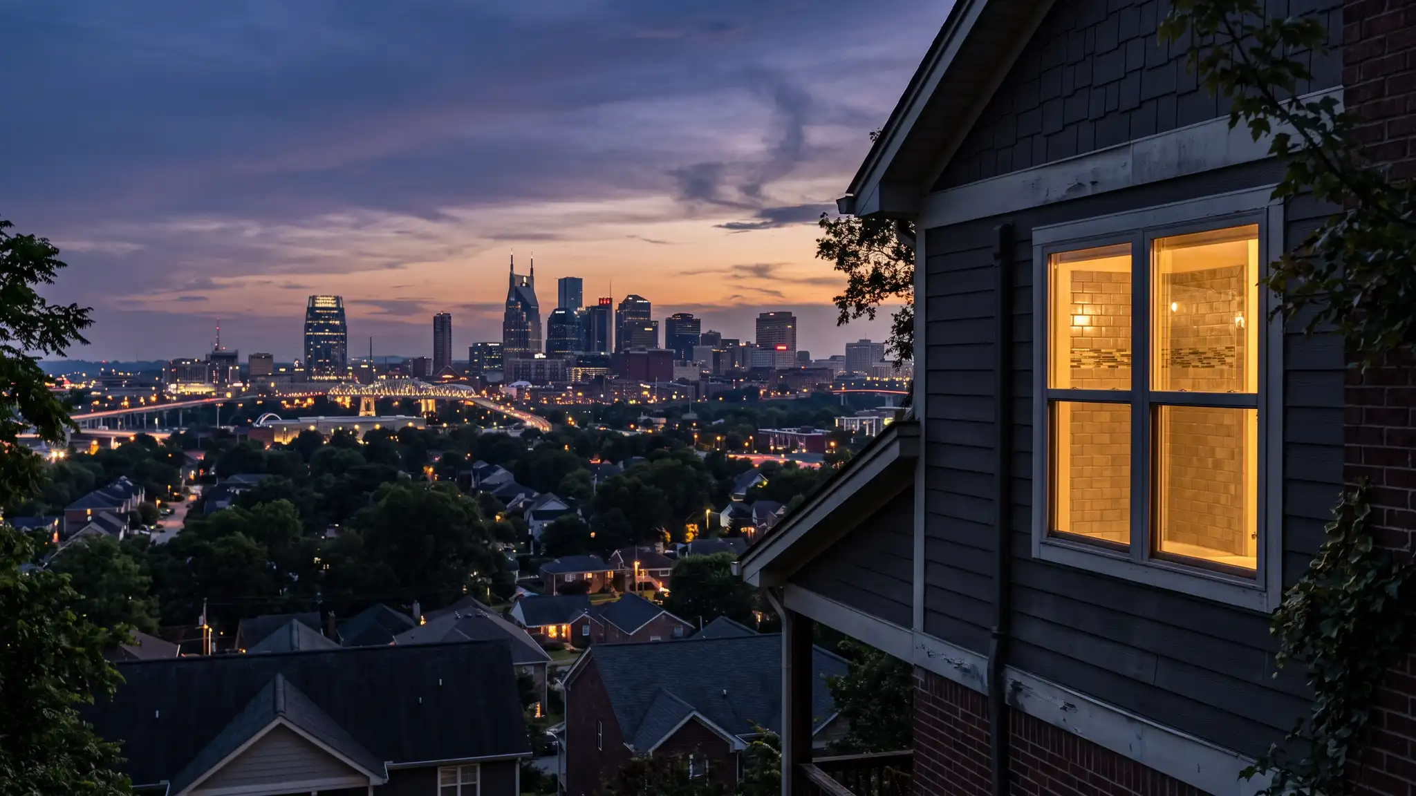 A dramatic dusk view of the Nashville skyline with warm lights over rooftops and a glowing tiled bathroom window of a suburban home in the foreground